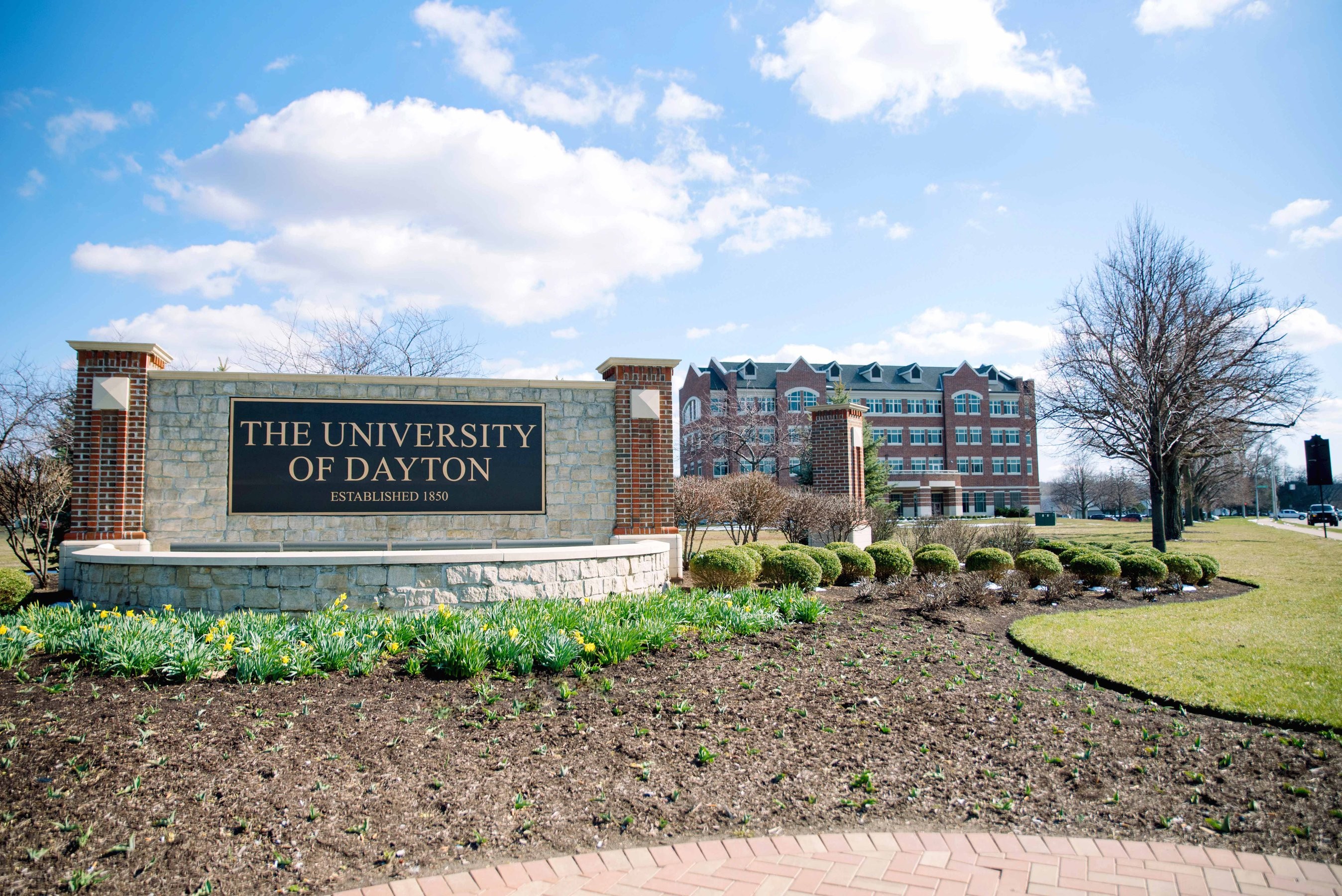 A sign for the University of Dayton in front of a campus building on a sunny day