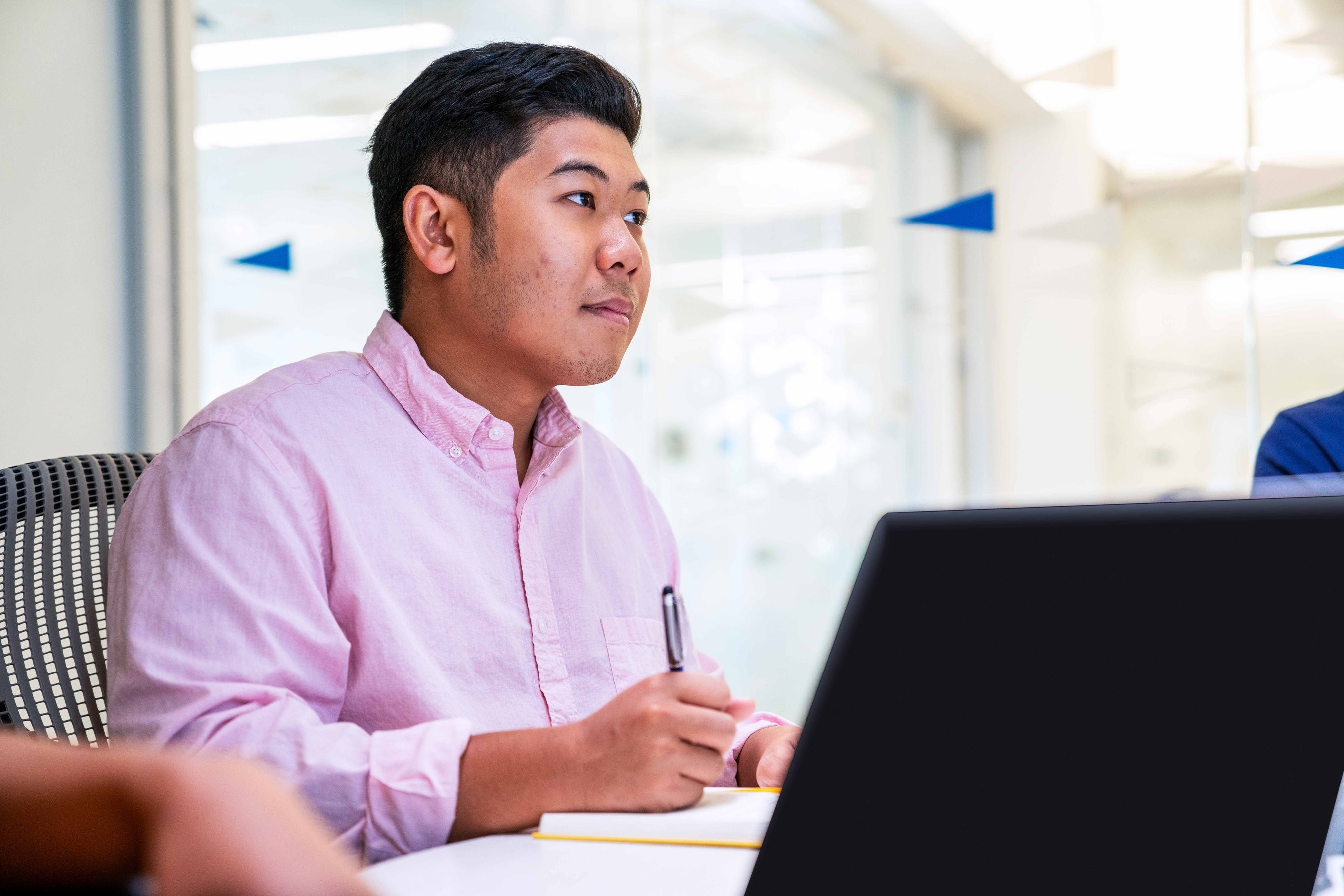 A young intern in a pink button-down shirt listens intently while taking notes at a meeting